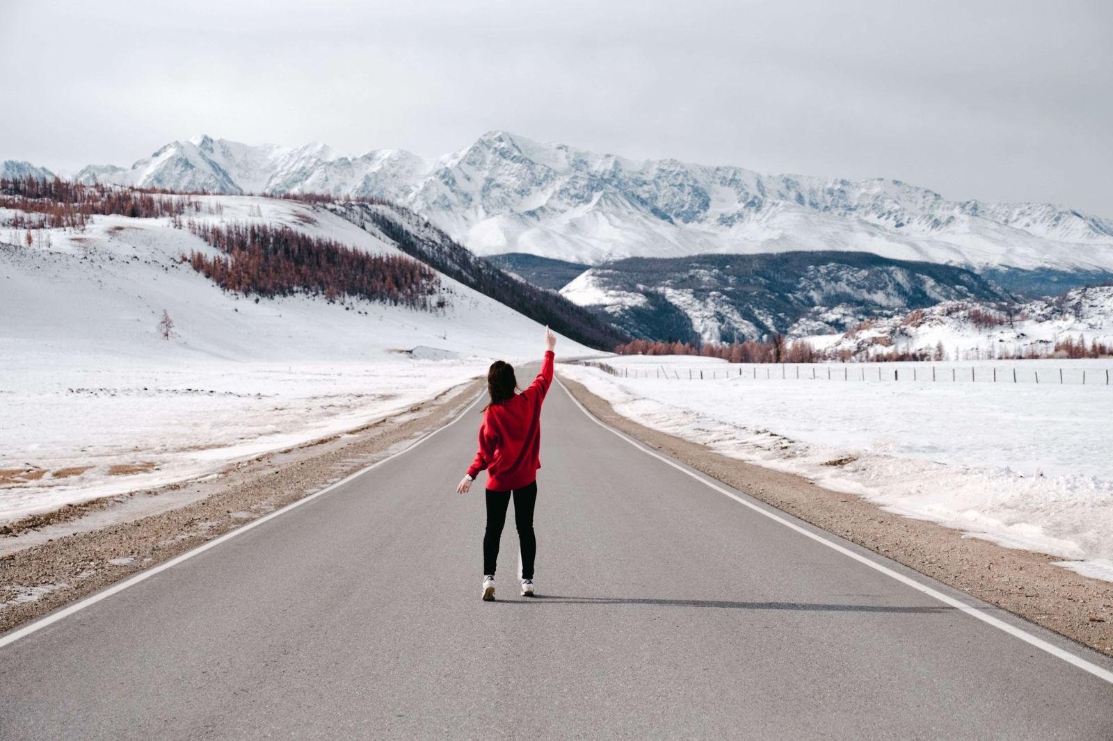 A woman walking along a long snowy road, one arm raised towards the mountains